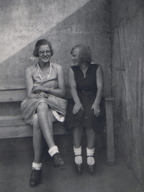 Zwei junge Frauen sitzen auf einer Bank. Die Frau links blickt in die Kamera, die rechts blickt auf die Frau links. Sie haben beide leichte Sommerkleider an.
